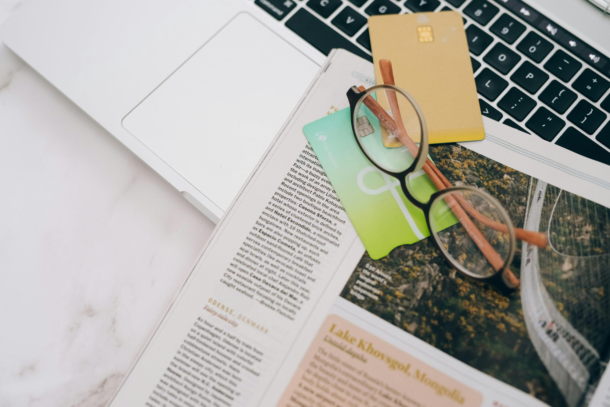 Travel credit cards and glasses on top of a travel magazine next to a laptop